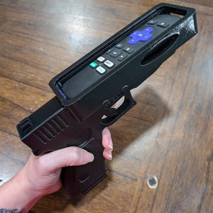 May include: A black 3D-printed remote control holder shaped like a handgun. The remote control is black with blue and white buttons. The holder has a trigger and grip, resembling a firearm. The image is taken from a slightly elevated angle.