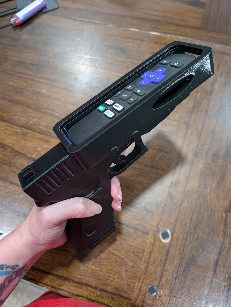May include: A black 3D-printed remote control holder shaped like a handgun. The remote control is black with blue and white buttons. The holder has a trigger and grip, resembling a firearm. The image is taken from a slightly elevated angle.