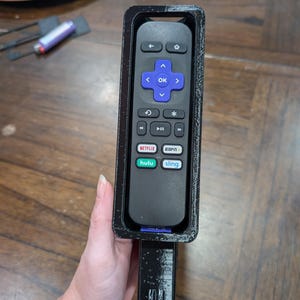 May include: A black Roku remote control inside a black 3D-printed holder. The remote has a directional pad with an "OK" button, and buttons for Netflix, Hulu, and Sling. The holder has a handle.
