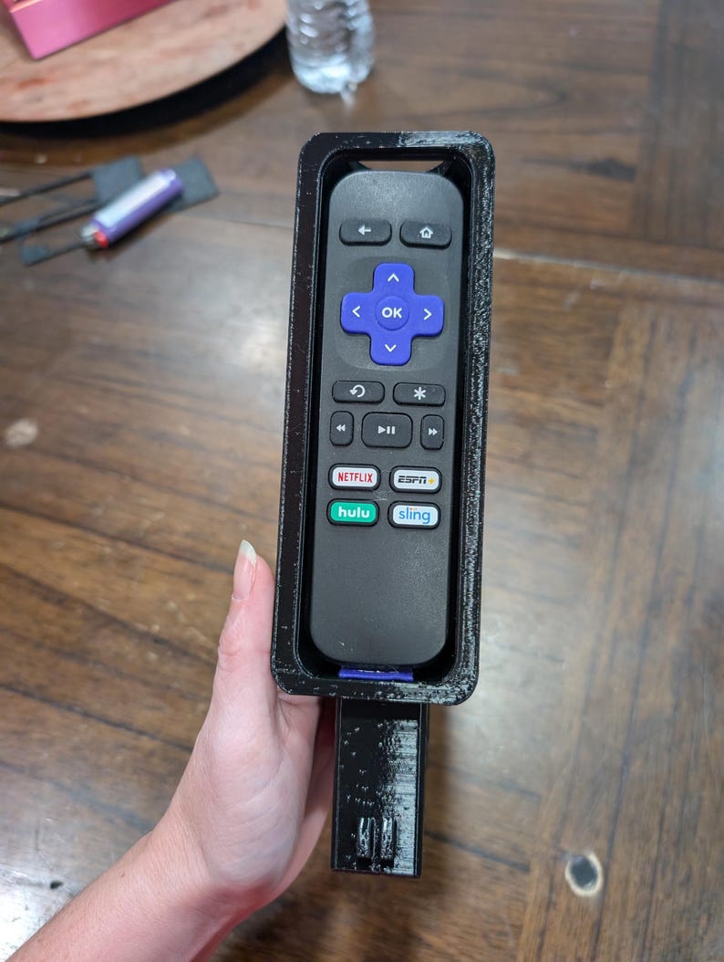 May include: A black Roku remote control inside a black 3D-printed holder. The remote has a directional pad with an "OK" button, and buttons for Netflix, Hulu, and Sling. The holder has a handle.