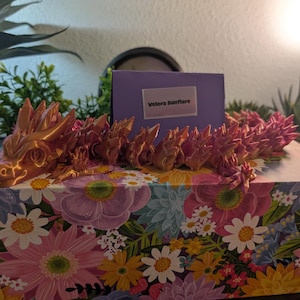 May include: A detailed, articulated dragon figurine in shades of copper and pink rests on a floral-patterned box. The dragon's head is prominently displayed, with intricate detailing. A purple card with the text "Velora Sunflare" is in the background.