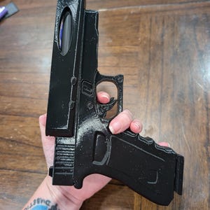 May include: A black 3D-printed replica handgun. The model features a detailed design, including the trigger, grip, and slide. The handgun is held against a wooden surface.