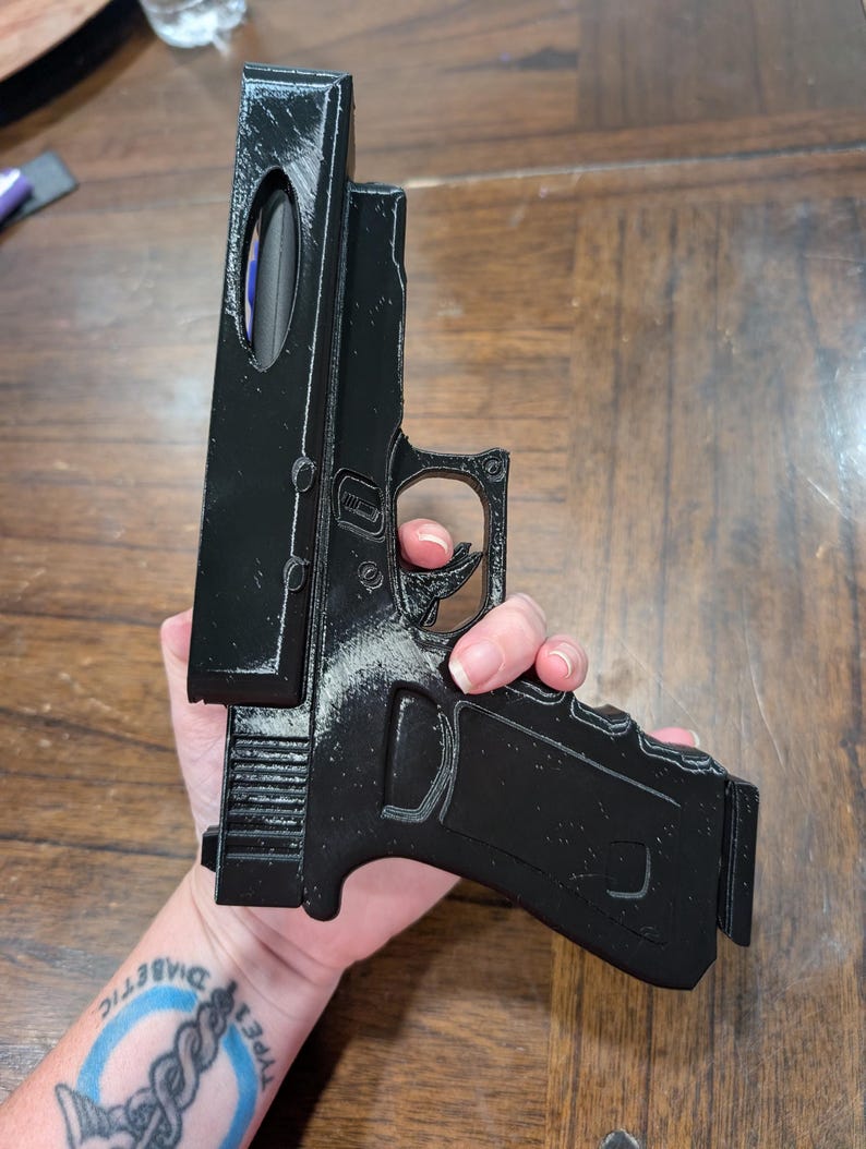 May include: A black 3D-printed replica handgun. The model features a detailed design, including the trigger, grip, and slide. The handgun is held against a wooden surface.