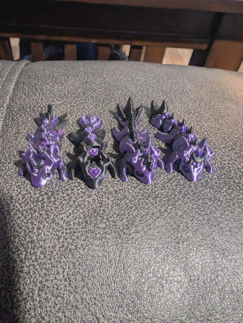 K&ouml;nnte beinhalten: Vier bewegliche Drachenfiguren in einer metallisch violetten und schwarzen Farbgebung. Jeder Drache verf&uuml;gt &uuml;ber einen segmentierten K&ouml;rper, einen detaillierten Kopf mit H&ouml;rnern und einen Schwanz. Die Figuren sind in einer Reihe angeordnet.