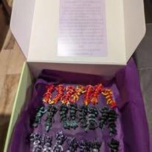 K&ouml;nnte beinhalten: Ein wei&szlig;er Karton mit einer Sammlung kleiner, beweglicher Kunststofffiguren, die in drei Reihen angeordnet sind. Die oberste Reihe enth&auml;lt rote Figuren, die mittlere Reihe dunkelgr&uuml;ne Figuren und die unterste Reihe besteht aus schwarzen Figuren.