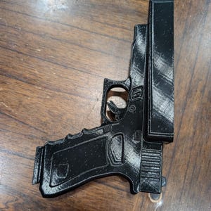 May include: A black 3D-printed replica of a handgun, featuring a detailed design of the frame, slide, and magazine. The gun is positioned on a wooden surface, showcasing its form and texture.