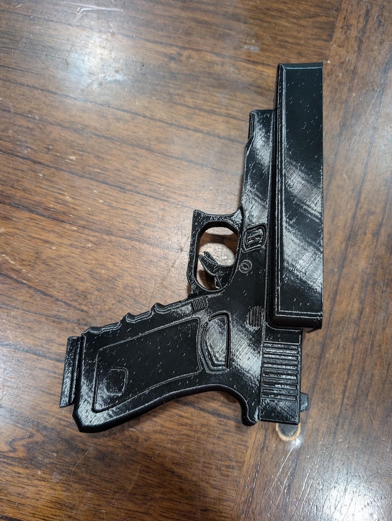May include: A black 3D-printed replica of a handgun, featuring a detailed design of the frame, slide, and magazine. The gun is positioned on a wooden surface, showcasing its form and texture.