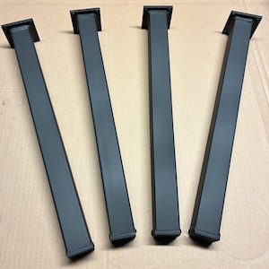 May include: Four black metal table legs with a square top and bottom. The legs are rectangular and have a matte finish. The legs are angled slightly and are arranged in a row.