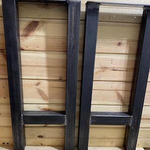 May include: Two rectangular table legs constructed from dark grey metal tubing. Each leg features two vertical posts connected by a horizontal crossbar near the bottom, with a flat mounting plate at the top. The metal surface shows visible weld marks.