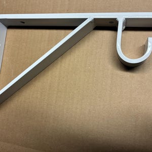 May include: A white metal L-shaped wall bracket featuring a diagonal support brace and a circular hook attachment at the top end. The bracket includes pre-drilled circular holes along the vertical and horizontal sections for mounting purposes.