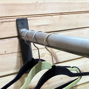 May include: A metal clothing rack mounted on a wooden wall. The rack has a cylindrical bar with a black end cap and a metal bracket. Several velvet hangers in black and light green hang from the bar.