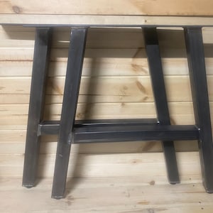 May include: Two black metal table legs with a rectangular top bar. The legs are made of thick metal and have a simple, industrial design.