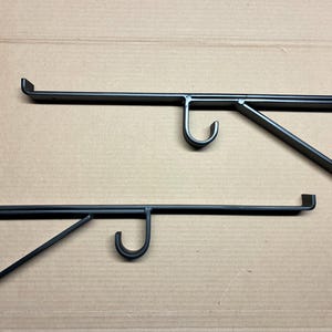 Pode incluir: Duas prateleiras de metal preto com design de gancho. Os suportes têm formato de ângulo reto com uma placa de montagem vertical e um braço horizontal. O braço apresenta um gancho curvo para pendurar objetos.