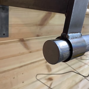 May include: Close-up of a metal pipe with a round end cap. The pipe is attached to a metal bracket and is mounted to a wooden wall.