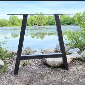 May include: A dark gray metal table leg frame featuring a trapezoidal shape with two angled vertical supports connected by a horizontal crossbar at the base and a flat mounting plate at the top, constructed from sturdy industrial steel tubing.