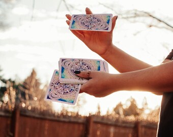 Könnte beinhalten: Die Hände einer Person halten einen Stapel Tarotkarten mit einem bunten Kompassdesign. Die Karten werden aufgefächert, wobei eine Karte in der Luft schwebt.