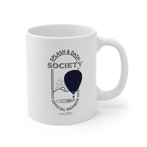 Könnte beinhalten: Weiße Keramiktasse mit einem schwarz-weißen grafischen Design. Das Design zeigt einen Heißluftballon, eine Sonne und den Text "Splash & Dash Society Official Member - Since 2015."