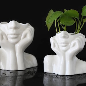 May include: Two white ceramic face planters, one empty and one with green leafy plants. The planters have a human face design with hands supporting the chin. The background is black.