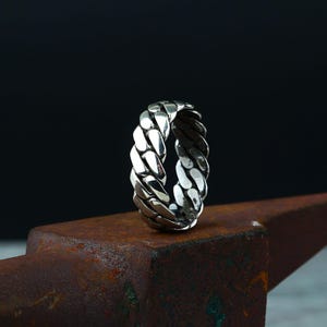 May include: A silver ring with a woven chain design. The ring is made of shiny metal and has a textured surface. The ring is displayed on a rusty metal surface against a dark background.