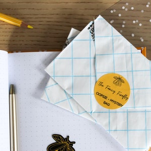 Puede incluir: Un sobre blanco con un patrón de cuadrícula azul y una pegatina circular amarilla que dice "The Fancy Firefly OOPSIE - MYSTERY BAG" con una ilustración de luciérnaga negra y amarilla. Una pegatina de luciérnaga negra y amarilla está en una libreta blanca con un patrón de puntos.