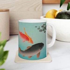 Könnte beinhalten: Weiße Keramiktasse mit blauem Hintergrund und einem Bild von drei Fischen, die im Wasser schwimmen. Die Fische sind orange, gelb und grau. Im Hintergrund befinden sich grüne Seerosen.