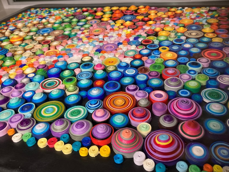 May include: A colorful mosaic art piece made of rolled paper circles. The circles are arranged in a circular pattern, creating a swirling effect. The colors are vibrant and varied, including red, orange, yellow, green, blue, purple, and pink.