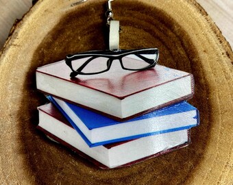 May include: A pair of earrings featuring a stack of three miniature books with red and blue covers, topped with a pair of black-framed glasses. The earrings are suspended from silver-colored fish hook ear wires.