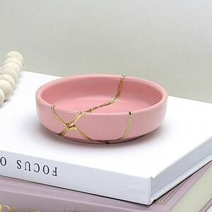 May include: A pink ceramic trinket dish with gold crackle detailing sits on top of two books. A white beaded garland with a brown tassel hangs off the edge of the books. A white vase with a pink rose and white baby's breath sits in the background.