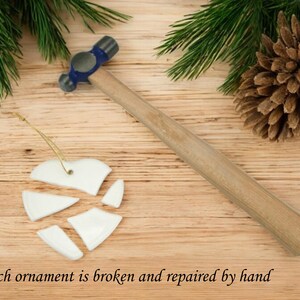 Peut inclure: Une d&eacute;coration de No&euml;l en forme de c&oelig;ur en c&eacute;ramique blanche bris&eacute;e en morceaux et un marteau bleu et argent&eacute; sur une surface en bois. Chaque d&eacute;coration est bris&eacute;e et r&eacute;par&eacute;e &agrave; la main.