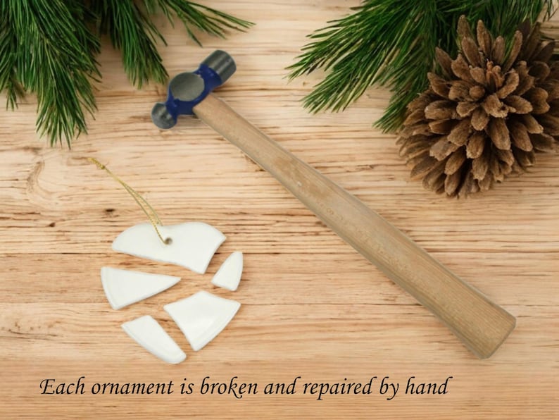 Peut inclure: Une d&eacute;coration de No&euml;l en forme de c&oelig;ur en c&eacute;ramique blanche bris&eacute;e en morceaux et un marteau bleu et argent&eacute; sur une surface en bois. Chaque d&eacute;coration est bris&eacute;e et r&eacute;par&eacute;e &agrave; la main.