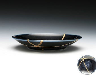 May include: A black ceramic plate with gold cracks. The plate is slightly curved and has a blue rim.