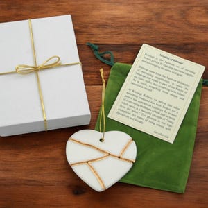 Peut inclure: Ornement en forme de c&oelig;ur en c&eacute;ramique blanche avec des accents dor&eacute;s, emball&eacute; dans une bo&icirc;te cadeau blanche avec un ruban dor&eacute; et un sac en velours vert. Le sac comprend une carte avec le texte "Signification de Kintsugi : Le Kintsugi est l'art japonais de r&eacute;parer la poterie cass&eacute;e en r&eacute;parant les morceaux ensemble et en accentuant les joints avec de l'or."