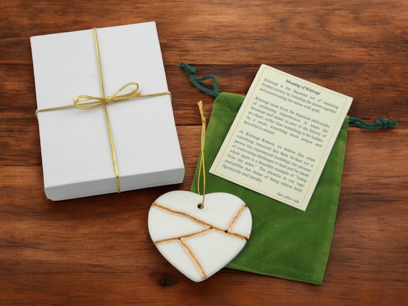Peut inclure: Ornement en forme de c&oelig;ur en c&eacute;ramique blanche avec des accents dor&eacute;s, emball&eacute; dans une bo&icirc;te cadeau blanche avec un ruban dor&eacute; et un sac en velours vert. Le sac comprend une carte avec le texte "Signification de Kintsugi : Le Kintsugi est l'art japonais de r&eacute;parer la poterie cass&eacute;e en r&eacute;parant les morceaux ensemble et en accentuant les joints avec de l'or."