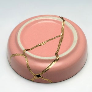 May include: A pink ceramic bowl with a crackled finish and gold accents. The bowl is cracked and repaired with gold kintsugi.