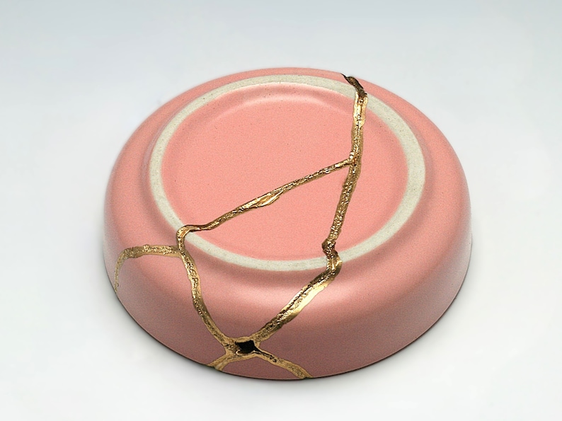 May include: A pink ceramic bowl with a crackled finish and gold accents. The bowl is cracked and repaired with gold kintsugi.