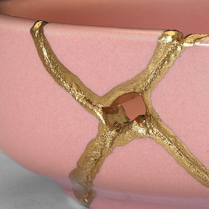 May include: A pink ceramic bowl with gold crackle glaze. The bowl has a unique design with gold lines that create a criss-cross pattern.