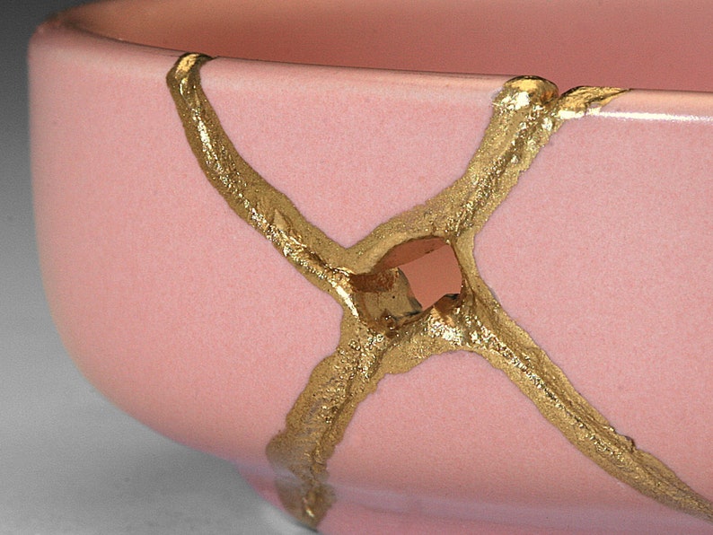 May include: A pink ceramic bowl with gold crackle glaze. The bowl has a unique design with gold lines that create a criss-cross pattern.