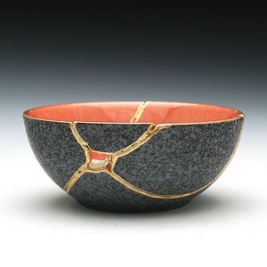 May include: A black ceramic bowl with a speckled texture, featuring gold-colored lines that mend cracks. The interior of the bowl is a vibrant coral color. This piece showcases the Japanese art of Kintsugi.