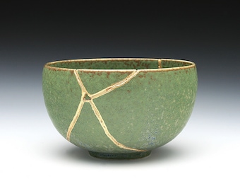May include: A green ceramic bowl, repaired using the Kintsugi technique, with gold-coloured lines. The bowl has a speckled texture and a rounded shape, with a brown rim. The gold lines highlight the repaired cracks.