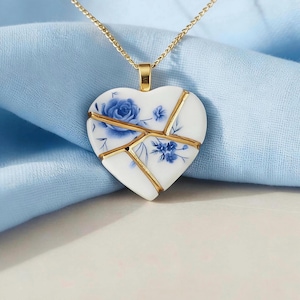 May include: A heart-shaped pendant necklace featuring a white ceramic base with blue floral patterns. The heart is divided into segments by gold-colored lines, mimicking the kintsugi technique, and hangs from a thin gold-colored metal chain.