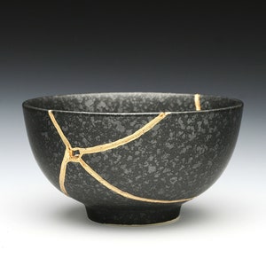 May include: A black ceramic bowl with a speckled texture, repaired with gold-colored lacquer using the Kintsugi method. The bowl has a round shape and a small foot, showcasing the beauty of imperfection.