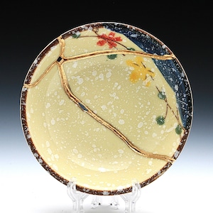 May include: A yellow ceramic plate with a blue and brown rim. The plate has a crack that is filled with gold paint. The plate is decorated with flowers and leaves.