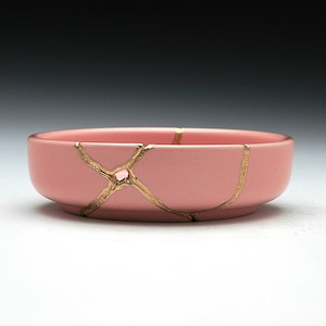 May include: A pink ceramic bowl with gold cracks. The bowl is round and has a smooth surface.