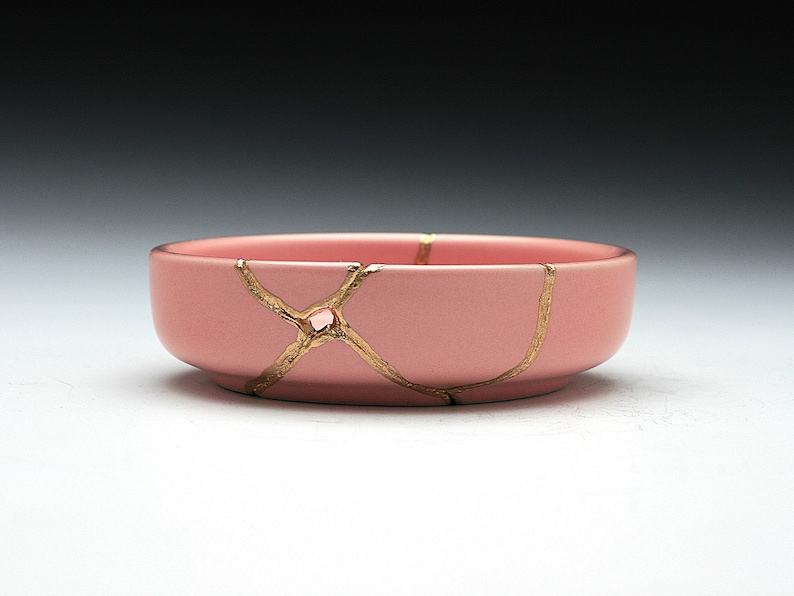 May include: A pink ceramic bowl with gold cracks. The bowl is round and has a smooth surface.
