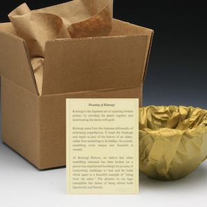 May include: A brown cardboard box with a gold tissue paper-wrapped bowl inside. The box is open and a card with the text "Meaning of Kintsugi" is visible. The card explains the Japanese art of repairing broken pottery with gold, called Kintsugi.