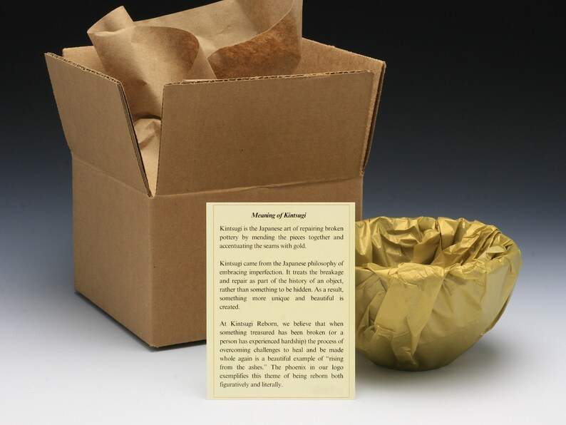 May include: A brown cardboard box with a gold tissue paper-wrapped bowl inside. The box is open and a card with the text "Meaning of Kintsugi" is visible. The card explains the Japanese art of repairing broken pottery with gold, called Kintsugi.