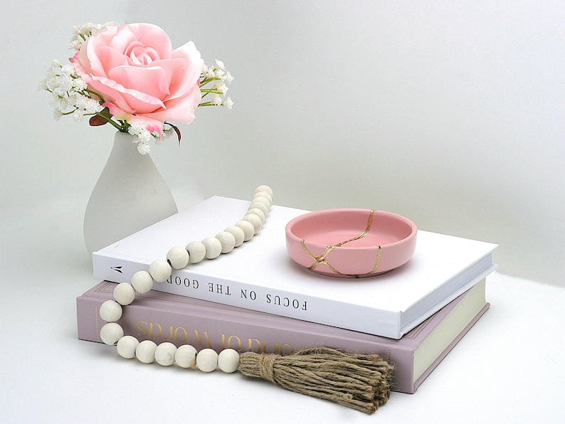 May include: A pink ceramic trinket dish with gold crackle detailing sits on top of two books. A white beaded garland with a brown tassel hangs off the edge of the books. A white vase with a pink rose and white baby's breath sits in the background.