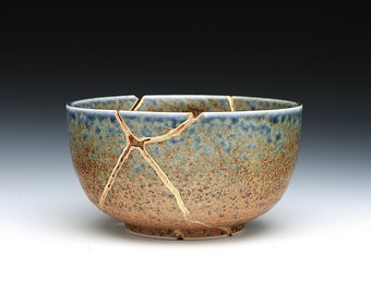 May include: A ceramic bowl with a speckled brown and blue glaze, repaired with gold-colored kintsugi. The bowl has a wide, open shape and a slightly flared rim. The gold lines highlight the cracks.