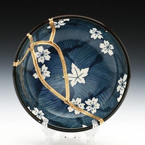 May include: A round, blue ceramic bowl with white floral and leaf designs. The bowl has been repaired with gold-colored kintsugi, a Japanese art form. The rim is black, and the bowl is displayed on a clear stand.