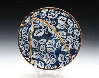 May include: A blue and white ceramic plate with a floral pattern and gold cracks. The plate is on a clear stand.